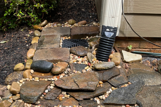 drain-5 Rain gutter drains into a black grate surrounded by rocks and stones near a house corner.