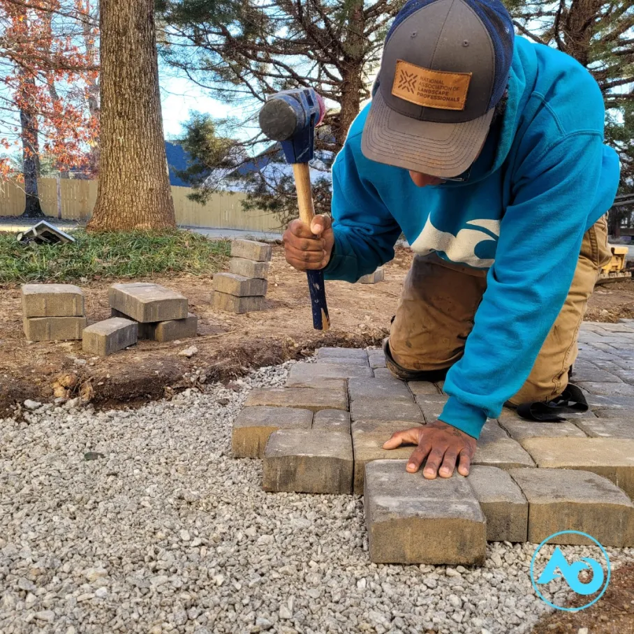 hardscape-2 A person kneels, laying paving stones and tapping them with a mallet on a gravel surface outdoors.