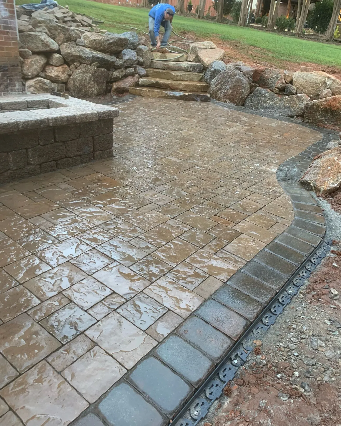 hardscape-benefits A person works on a wet stone patio, building steps and arranging rocks in a landscaped yard.
