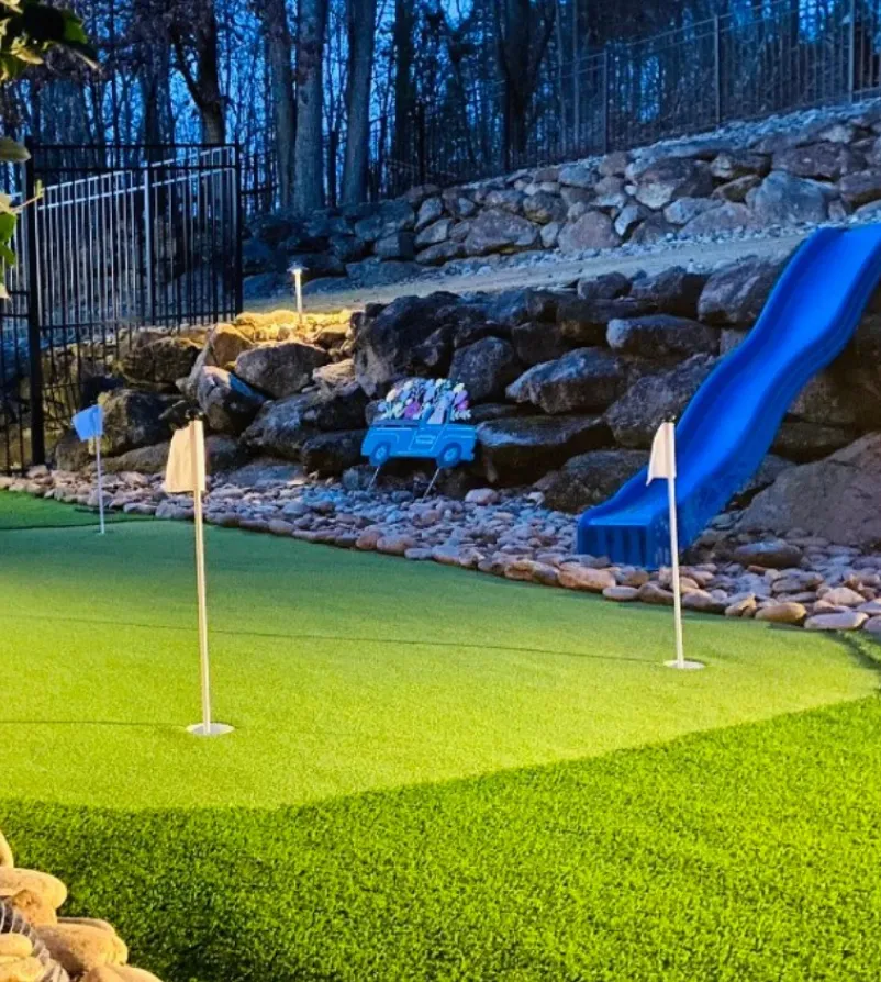 putting-green-1 (1) A backyard mini golf green with white flags, rocks, and a blue slide near a stone wall at dusk.