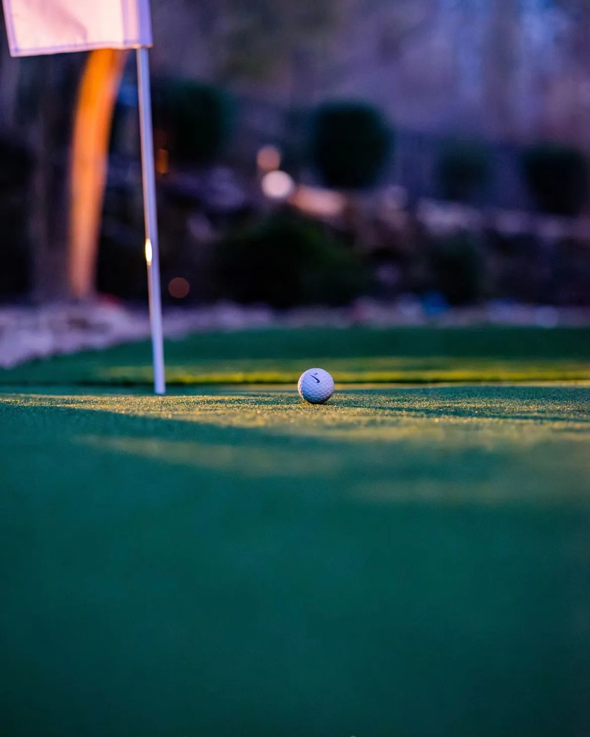 putting-green-3 A golf ball resting on green grass near a flagstick at sunset or dusk.