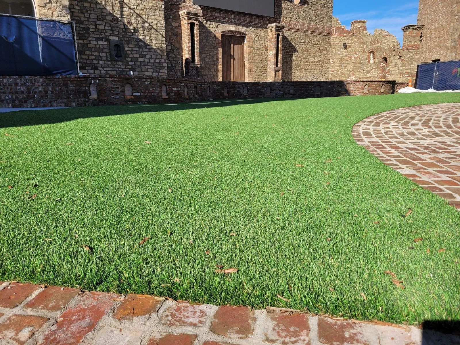 serv-h A patch of green grass beside a brick pathway and old stone building under a blue sky.
