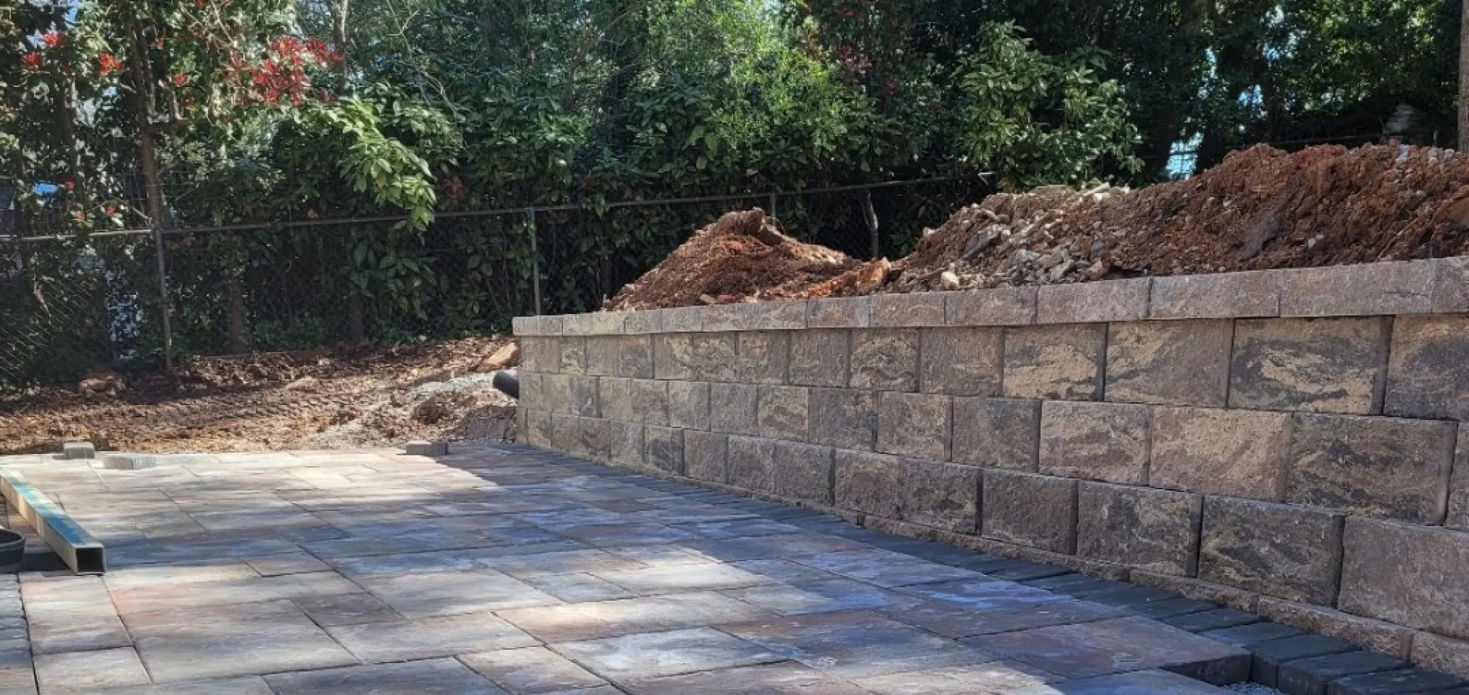 walls-4 A stone retaining wall with dirt behind it, next to a paved patio and surrounded by trees.
