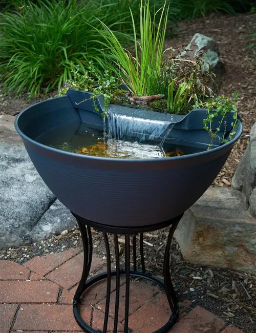 Large black bowl fountain with flowing water and green plants, set on a stand in a garden.